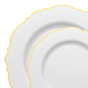 Two plates from the Kaya Collection's White with Gold Rim Round Blossom Disposable Plastic Dinnerware Value Set, featuring wavy edges and an elegant gold trim along the rims. The minimalist design is perfect for party use. The smaller plate is stacked on top of the larger one, showcasing their scalloped borders.