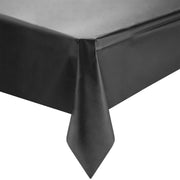 A close-up view of a Kaya Collection Black Rectangular Disposable Plastic Tablecloths (54" x 108") draped over the edge of a table. The fabric hangs down, creating a smooth, elegant appearance with a pointed corner. The texture of the tablecloth appears silky and slightly reflective, perfect for sleek party table decor.