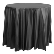 A round table covered with a Kaya Collection Black Round Disposable Plastic Tablecloths (84") that drapes down to the floor. The shiny, smooth material of the plastic tablecloth creates slight ripples and folds along the edges. The table stands alone against a white background.