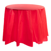 A round table is adorned with an 84-inch Red Round Disposable Plastic Tablecloth that extends to the floor. The vibrant red tablecloth has slight creases and folds, giving it a flowing appearance. The background is plain and white. This event table cover effortlessly elevates the entire setting.