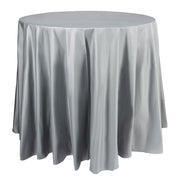 A round table covered with a shiny Silver Round Disposable Plastic Tablecloth (84") that drapes down to the floor. The silver party tablecloth features soft folds and a slightly wrinkled appearance, adding a subtle texture to its otherwise sleek surface.