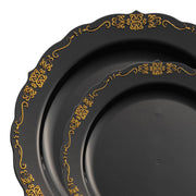 Two ornate plates from the Kaya Collection's Black with Gold Vintage Rim Round Disposable Plastic Dinnerware Value Set feature intricate gold floral designs on their edges. The elegant dinnerware boasts scalloped rims, with the larger plate partially overlapping the smaller one to create a layered effect. The gold detailing adds a sophisticated touch to the overall appearance.