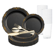 The Smarty had a party Black with Gold Vintage Rim Round Disposable Plastic Wedding Value Set includes large and small plates, forks, spoons, knives, and clear plastic cups stacked in the background. The elegant design features intricate gold patterns along the edges of the plates, making them ideal disposable party supplies.