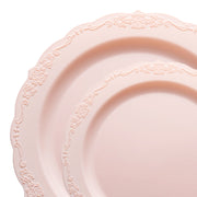 A close-up image of two stacked, round plates from the Pink Vintage Round Disposable Plastic Dinnerware Value Set by Kaya Collection, featuring light pink color with decorative floral patterns along their scalloped edges. The intricate design adds an elegant touch, perfect for a romantic wedding reception. The plates appear glossy and smooth.