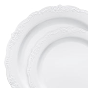 Two plates from the Kaya Collection's White with Silver Vintage Rim Round Disposable Plastic Dinnerware Value Set, featuring intricate embossed floral patterns along the rims. The partially overlapping plates showcase their elegant design and scalloped edges against a plain white background, offering an elevated touch compared to typical disposable dinnerware.