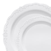 Close-up of two pieces from the Kaya Collection's White Vintage Round Disposable Plastic Dinnerware Value Set, featuring delicate embossed floral patterns along the rims. The plate in front is slightly smaller, overlapping the larger one behind it. Both plates have an elegant, ornamental design that perfectly complements any sophisticated table settings.