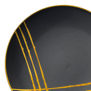 A close-up of a black disposable plastic plate from the Kaya Collection, boasting a modern design with gold brushstrokes. Four parallel gold lines run vertically from the left edge towards the center, with another intersecting horizontally near the bottom left corner, reminiscent of elegant Black with Gold Brushstroke Round Disposable Plastic Dinner Plates (10.25").