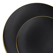 A close-up image showcases two stacked, round black plates with delicate gold-rimmed borders from the Kaya Collection's Black with Gold Rim Organic Round Disposable Plastic Dinnerware Value Set. The larger plate sits beneath the smaller one, both partially visible within the white background, which accentuates the striking contrast of the black and gold in this elegant disposable dinnerware set.