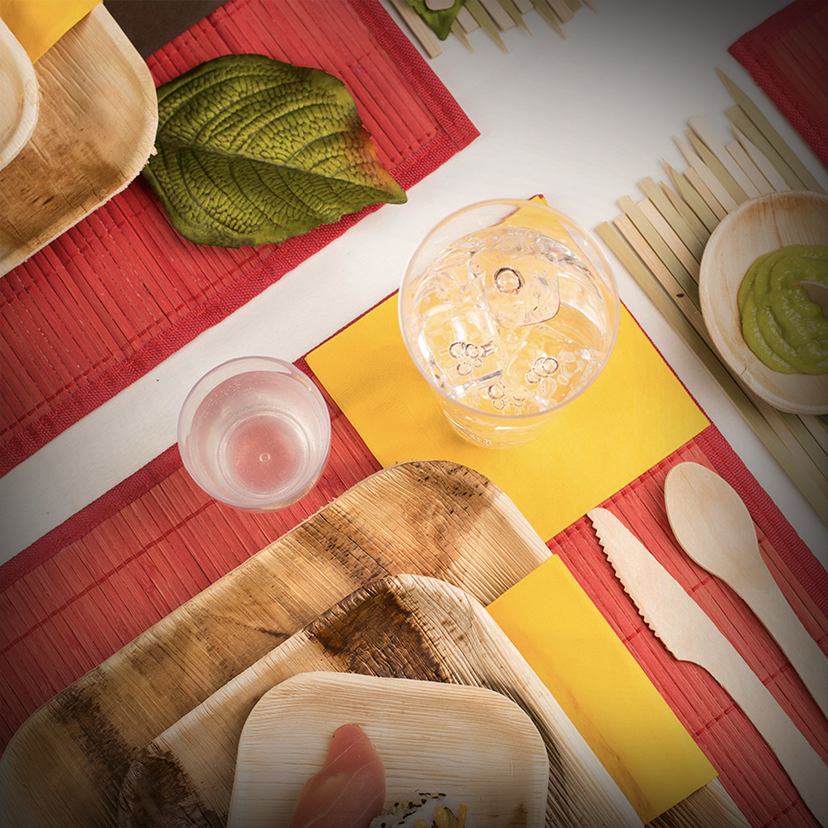 A top view of a table setting with eco-friendly bamboo plates and cutlery on red and yellow place mats. The table also has a water glass, a smaller glass with a clear beverage, a dish of green sauce, and a leaf decoration. A sashimi piece is on one of the plates.