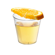 A 1 oz. Clear Plastic Shot Glass from Kaya Collection, filled with a light yellow liquid and topped with a large slice of orange, set against a clean, white surface.