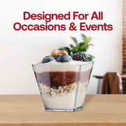 A stylish square cup, specifically the 3.5 oz. Clear Square Disposable Plastic Mini Cup with Lid, containing layers of granola, yogurt, fruit puree, and topped with blueberries and blackberries sits on a wooden table. The translucent clear design showcases the dessert beautifully. A blurred background features a plant and the text "Designed For All Occasions & Events" in large, red font.
