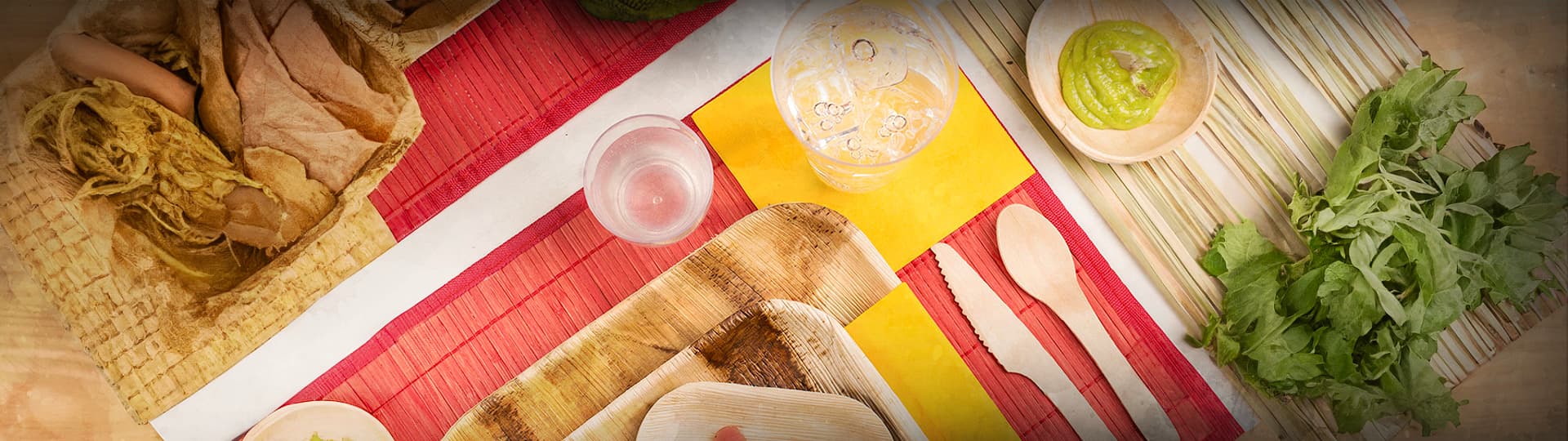 A variety of food items and utensils are laid out on a tabletop with colorful placemats. The items include bread, a glass of water, a wooden spoon and fork, a bowl of green dip, and fresh greens. The scene captures a vibrant and inviting meal setup.
